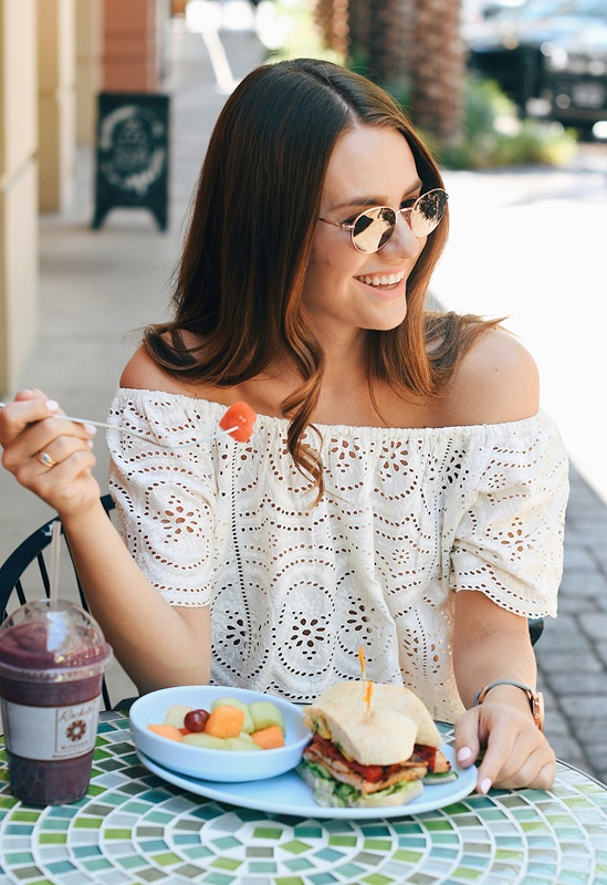 Woman sitting on a terrace, looking happy while eating a club sandwich with a blueberry smoothie